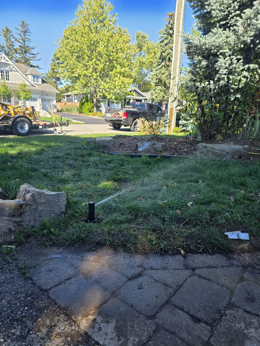 Pop-up sprinkler head watering a front lawn with the excavator and truck parked at the curb